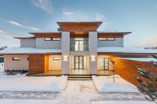 Primarily white house with wood accents around the house in the center ofthe photo with snow and a side walk.
