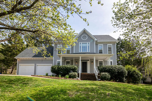 A large grey and white house with two garage doors and a front yard with green grass, bushes and some trees.